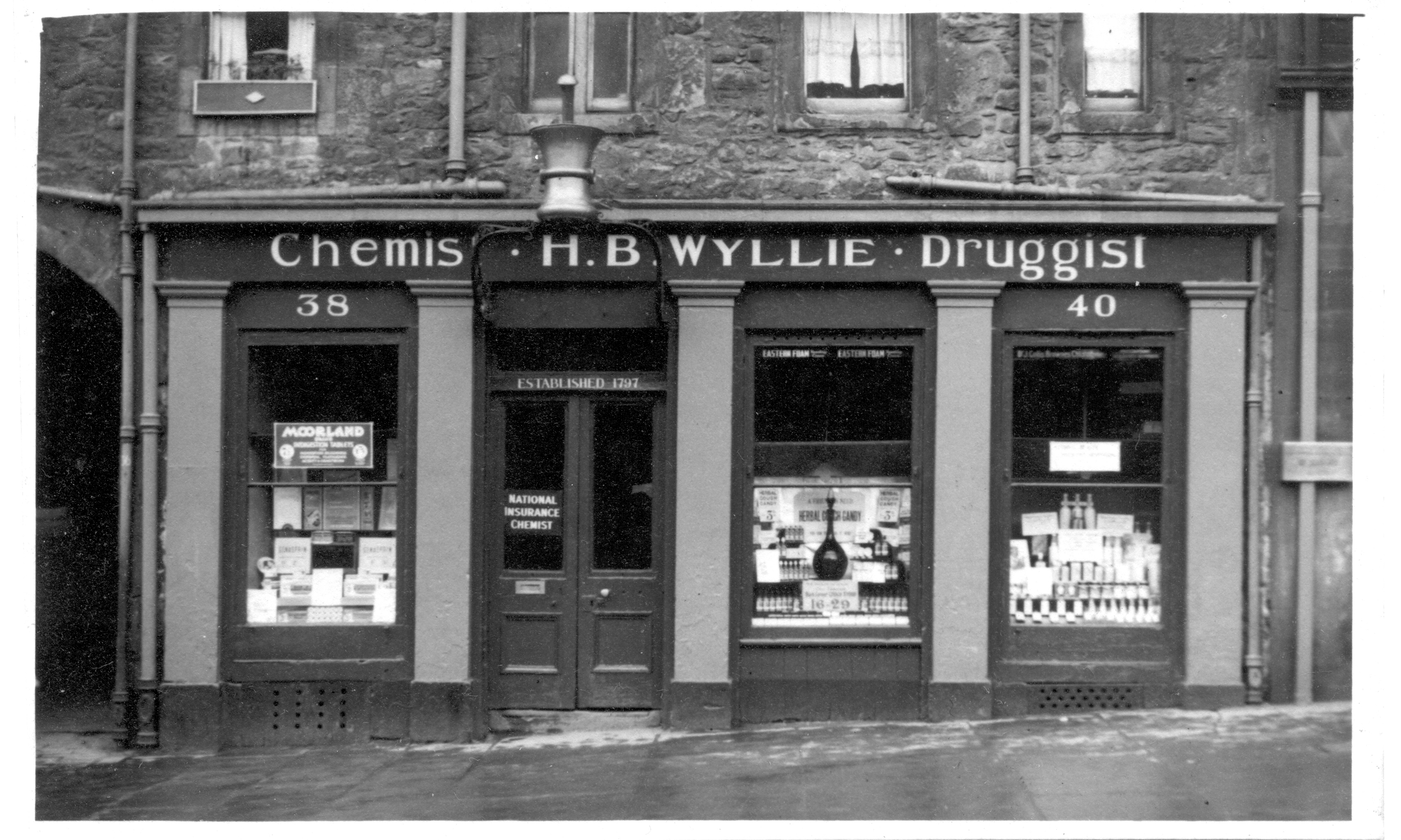 LDRPS: SZ3086, 1953. This black and white photograph shows the exterior of H.B Wyllie, a chemist and druggist in Edinburgh. The business was established in 1797
